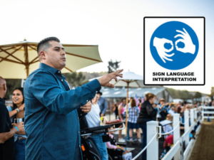 A man is providing sign language interpretation at an outdoor event, ensuring accessibility for attendees who are deaf or hard of hearing. He is wearing a blue shirt and is actively signing while standing near a crowd of people who are engaged in the event. Behind him, there are umbrellas and a fenced area, creating a lively and inclusive atmosphere. A sign with the words "Sign Language Interpretation" and a symbol of hands signing is visible, highlighting the commitment to accessibility and inclusivity at The Shea Center.
