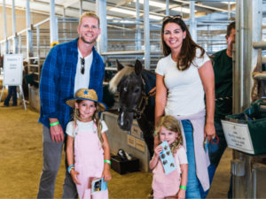 A family of four stands in a barn area next to a horse. The parents are smiling, and the two young girls, dressed in matching pink overalls, are excitedly posing with the horse. One girl is holding a booklet, and the other is wearing a straw hat. The setting is lively and engaging, reflecting a fun and educational experience at The Shea Center.
