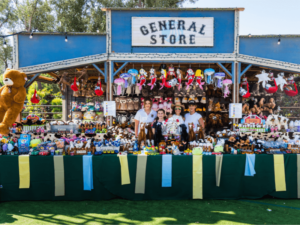 A colorful "General Store" booth at an outdoor event is filled with a variety of plush toys and other merchandise. Three staff members are standing behind the counter, smiling and ready to assist visitors. The booth is decorated with hanging toys and festive banners, creating a lively and inviting atmosphere. This vibrant scene showcases a fun and engaging part of The Shea Center's event, emphasizing the family-friendly and interactive experiences available.