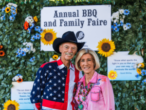 A smiling couple dressed in festive western attire stands in front of a sign that reads "Annual BBQ and Family Faire." The man is wearing a cowboy hat and a shirt with an American flag design, while the woman is wearing a pink shirt with decorative jewelry. The backdrop is decorated with sunflowers and greenery, creating a vibrant and cheerful atmosphere. This image captures the joyous spirit of the event, highlighting the community and family-oriented activities at The Shea Center's annual celebration.