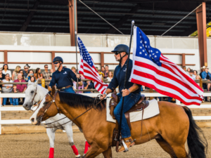 Two riders on horseback carry large American flags during a performance in an indoor arena. Both riders are dressed in dark shirts and helmets, and the horses are well-groomed and outfitted with saddles and reins. A white horse and a brown horse are shown, and the background features a crowd of spectators watching the event. This patriotic display emphasizes the spirit of community and the equestrian expertise at The Shea Center.