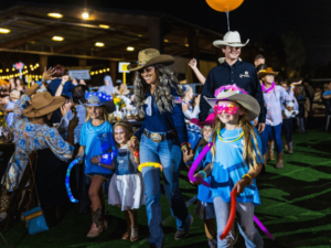 A group of children and adults participate in a lively nighttime event. The children, dressed in blue shirts and cowboy hats, are adorned with glow sticks and colorful accessories. The adults, also in cowboy hats, guide the children through the festivities. The event is well-lit with string lights, and a crowd is visible in the background, creating a vibrant and joyful atmosphere. This scene highlights the fun and engaging activities provided by The Shea Center, emphasizing their commitment to creating memorable experiences for all attendees.