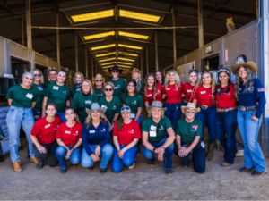 A group of staff and volunteers pose for a group photo inside a stable at The Shea Center. They are wearing coordinated outfits, with some in green shirts and others in red shirts, paired with jeans and cowboy hats. The group, consisting of around twenty people, smiles warmly at the camera, reflecting the camaraderie and teamwork within The Shea Center's community. The stable setting emphasizes the equestrian focus of their work, highlighting the dedication and collaborative spirit of the team.