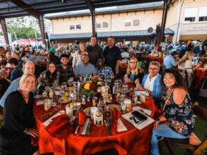 A group of people is seated around a large, round table covered with a red tablecloth at an indoor event. The table is set with plates, glasses, and festive decorations. The attendees are smiling and appear to be enjoying the gathering, which is part of a larger event with many other tables and people in the background. The setting is lively and filled with camaraderie, showcasing the sense of community and celebration at The Shea Center’s event.