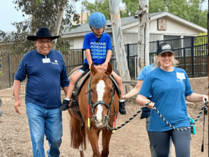 A smiling man in a black hat and a woman in a blue shirt and cap walk alongside a child riding a horse. The child, wearing a blue helmet and shirt, sits comfortably on the horse. They are outdoors on a dirt path, surrounded by trees and a building in the background. This scene captures the supportive and therapeutic environment of The Shea Center, highlighting the joy and engagement of the participants.