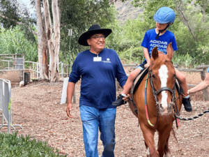 A smiling man in a blue shirt and black hat walks alongside a child riding a horse. The child is wearing a blue helmet and a blue shirt. They are outdoors on a dirt path with greenery and trees in the background. The scene exudes a sense of joy and therapeutic engagement, reflecting the supportive environment of The Shea Center's programs.