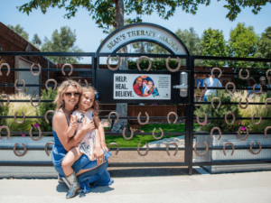 A smiling woman and a young girl stand in front of the entrance to Sean's Therapy Garden at The Shea Center. The gate, decorated with horseshoes, features a sign that reads "Help. Heal. Believe." The garden inside is lush and well-maintained, with greenery and flowers creating a serene atmosphere. The woman is wearing sunglasses and a blue dress, while the girl is in a floral dress and boots. They are both hugging and looking at the camera, radiating happiness and warmth, embodying the therapeutic and community spirit of The Shea Center.