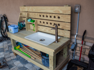 A wooden outdoor sink station designed for children's play and learning activities. The setup features a white sink with a faucet, a wooden backsplash with holes for storage, and various compartments and hooks for organizing tools and toys. The station is positioned on a tiled patio with additional play items and equipment nearby, providing a functional and interactive space for children to explore and engage in hands-on activities at The Shea Center.