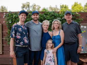 A group of six people, consisting of four adults and two children, are standing together and smiling in front of a wooden fence adorned with greenery. The adults are dressed in casual summer attire, and the children are wearing floral dresses. The scene depicts a warm, family-friendly environment, highlighting the supportive and community-oriented atmosphere of The Shea Center.