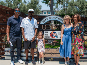 A group of five people, consisting of three adults, one child, and another adult, are standing in front of a gate that reads "Sean's Therapy Garden." The gate is adorned with horseshoes and a sign that says "Help. Heal. Believe." The adults are dressed in summer attire, and the young girl is wearing a floral dress. They all appear happy, enjoying a sunny day in a garden setting, emphasizing the welcoming and therapeutic environment of The Shea Center.