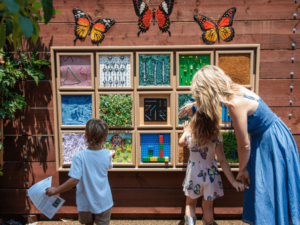 A woman and two young children are exploring a sensory wall in an outdoor garden. The wall features various textured panels designed for tactile interaction, adorned with decorative butterflies. The woman, dressed in a blue dress, is holding the hand of one child while the other child examines the panels. This engaging scene takes place in a vibrant and educational garden setting, emphasizing the interactive and supportive environment provided by The Shea Center.
