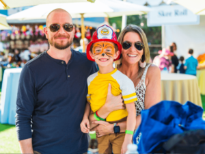 A smiling family of three enjoys a sunny day at an outdoor event. The father, wearing sunglasses and a dark shirt, stands next to the mother, also in sunglasses and a patterned dress. They are holding their child, who is dressed in a firefighter costume complete with a red helmet and face paint resembling a tiger. The background features festive booths and a lively crowd, highlighting the fun and family-friendly atmosphere of The Shea Center’s event.