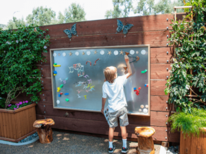 A young boy is engaged in drawing on a large outdoor magnetic whiteboard, which is mounted on a wooden fence adorned with decorative butterflies. The board is surrounded by lush greenery, planters, and tree stump seats, creating an inviting and interactive space. The boy is using colorful magnets and markers to create his artwork, highlighting the creative and educational activities available at The Shea Center.