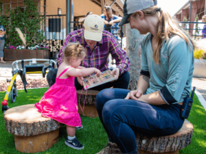A young girl in a pink dress plays with a wooden puzzle, assisted by two adults sitting on tree stump seats. The adults, one in a plaid shirt and hat, and the other in a gray shirt, are attentively engaging with the child, creating a nurturing and interactive environment. The scene takes place outdoors in a garden area with greenery and various play items in the background, highlighting the supportive and developmental activities offered at The Shea Center.