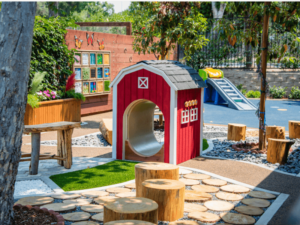 A vibrant outdoor therapy garden featuring a red playhouse shaped like a barn, surrounded by wooden pathways, tree stumps for seating, and lush greenery. The area includes sensory panels and educational displays on the wooden fence, adding a colorful and interactive element to the environment. In the background, there is a small playground slide, enhancing the engaging and therapeutic space designed to provide a supportive and stimulating experience for visitors at The Shea Center.