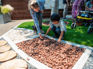A young boy is playing with a bed of smooth stones in an outdoor sensory garden, guided by an adult woman. They are engaged in a tactile activity, exploring the textures of the stones. The area is part of a larger garden with pathways, greenery, and other sensory elements, designed to provide therapeutic and developmental experiences. The environment is inviting and supportive, highlighting the interactive and nurturing activities offered at The Shea Center.