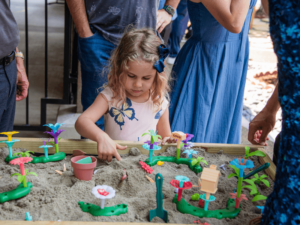 A young girl is deeply engaged in playing with a sand table filled with colorful toys and structures. She is focused on her activity, using her hands to explore and manipulate the items in the sand. Adults stand nearby, creating a supportive environment. The scene captures a moment of tactile play and exploration, emphasizing the engaging and developmental activities offered at The Shea Center.