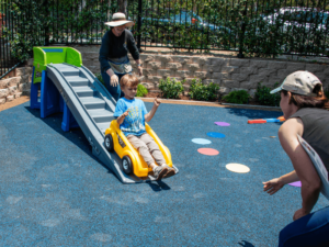 A child enjoys sliding down a small playground slide, with an adult assisting at the top and another adult waiting at the bottom. The playground surface is covered with a blue soft mat, and colorful circular markers are placed on the ground for additional play activities. The scene takes place in an outdoor area surrounded by a fence and greenery, highlighting the fun and safe environment provided for clients at The Shea Center.