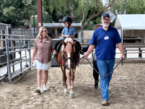 A child wearing a helmet rides a horse while being led by two volunteers, one on each side. The boy smiles confidently as the team walks him through an outdoor arena at The Shea Center. The volunteers offer support and guidance, ensuring a safe and enjoyable experience. The background features a mix of trees and stables, creating a calm and welcoming environment.