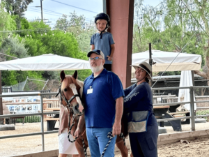 A young rider sits atop a horse, accompanied by two adults and a handler leading the horse. The handler, wearing a blue shirt and glasses, holds the horse's reins while standing beside the horse. Another individual, dressed in a wide-brimmed hat and casual attire, supports the rider, while a third person, partly visible, stands near the horse's side. They are in an outdoor covered area with trees and horse stables in the background. The rider is smiling, wearing a helmet, and enjoying the experience.
