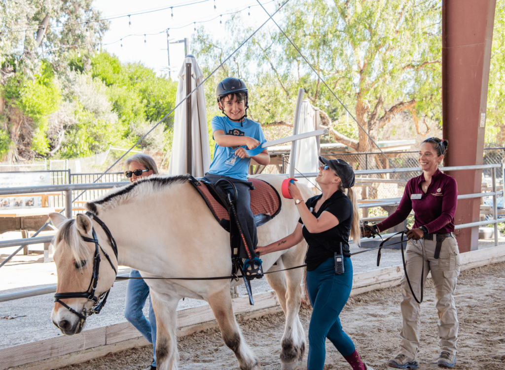 Child rider participates in a therapeutic session at The Shea Center, guided by supportive staff and volunteers. The rider is smiling while holding the reins, with a gentle horse walking in a covered arena.
